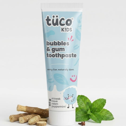Tube of tüco Kids bubbles & gum toothpaste with mint leaves and wooden sticks on a white background