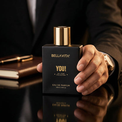 Hand holding a black Bellavita perfume bottle with gold cap on a reflective surface.