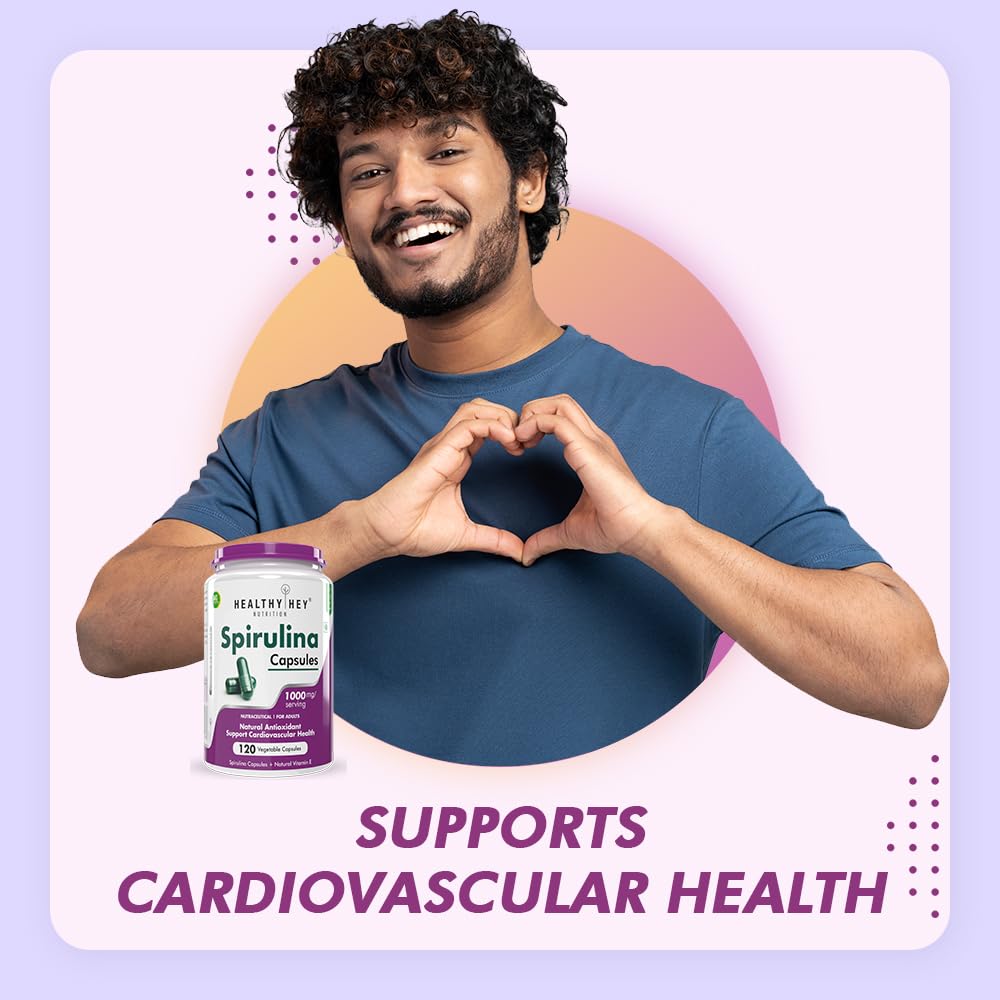 Man making a heart shape with his hands holding a bottle of Spirulina capsules against a light purple background.