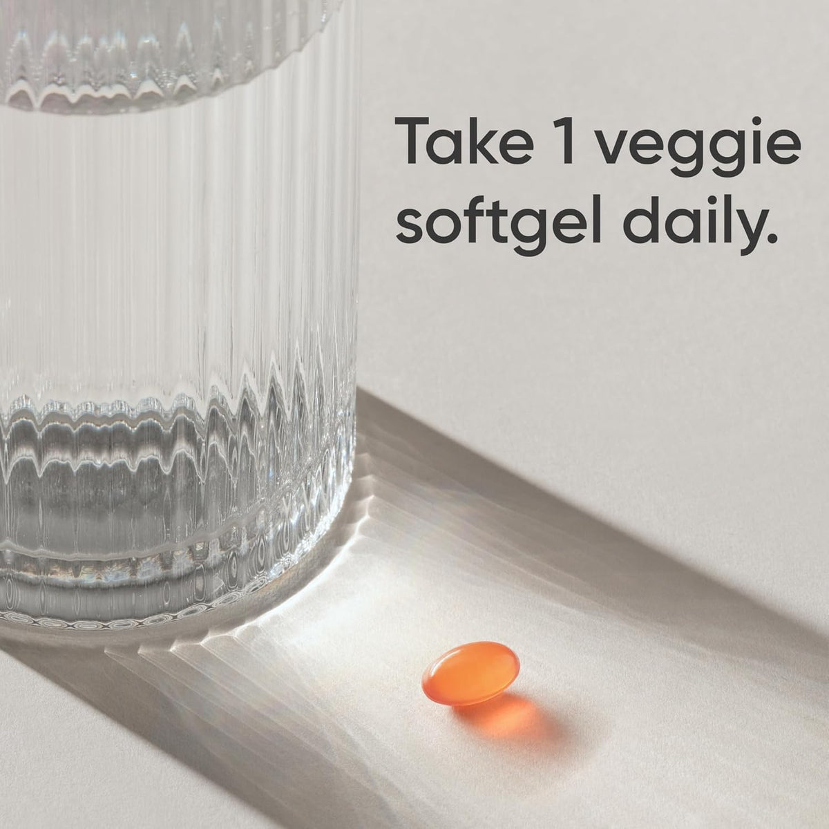 Orange pill next to a glass on a light surface with text 'Take 1 veggie softgel daily'.