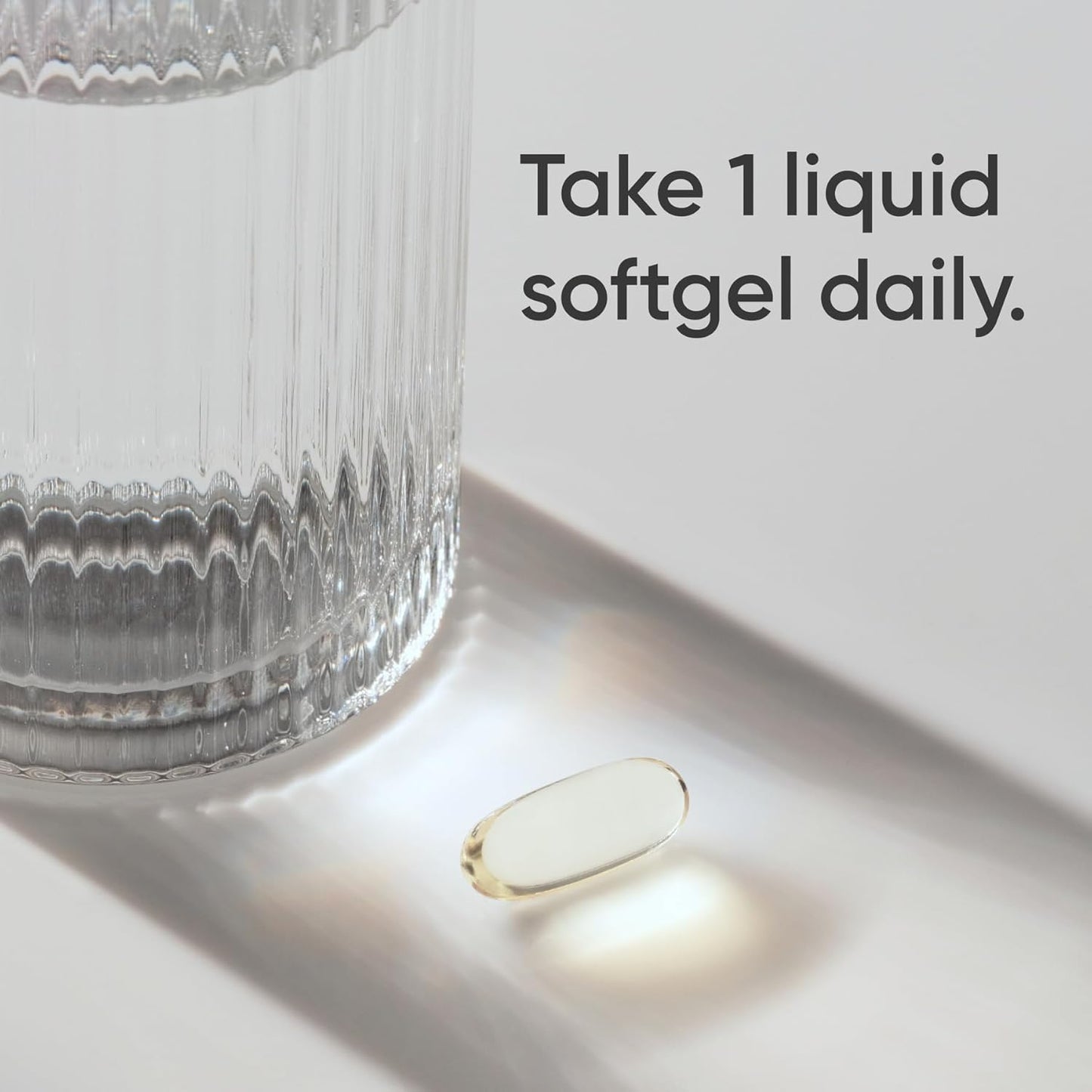 Vitamin supplement on a white surface with a glass of water and text 'Take 1 liquid softgel daily.'