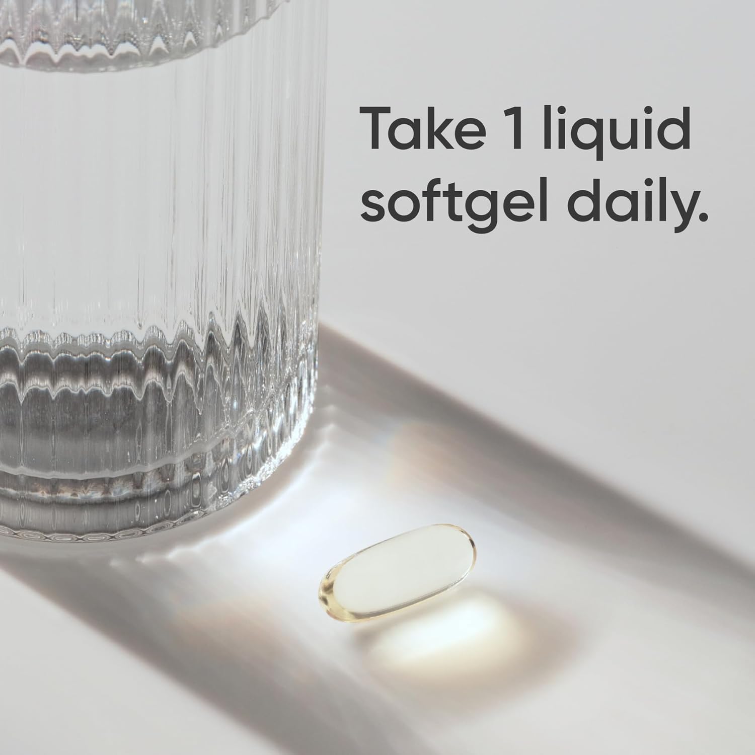 Vitamin supplement on a white surface with a glass of water and text 'Take 1 liquid softgel daily.'