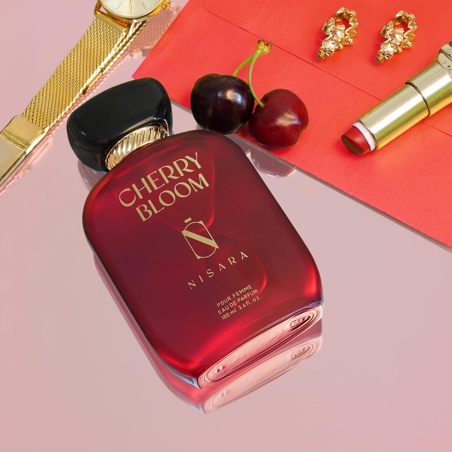 Red 'Cherry Bloom' perfume bottle by Nisara with cherries, lipstick, and watch on a pink background