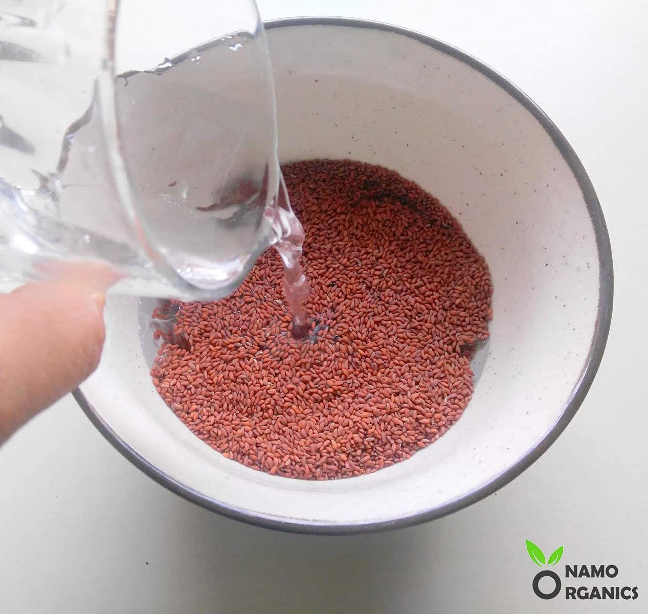 Person pouring water into a bowl of red seeds with Namo Organics logo.