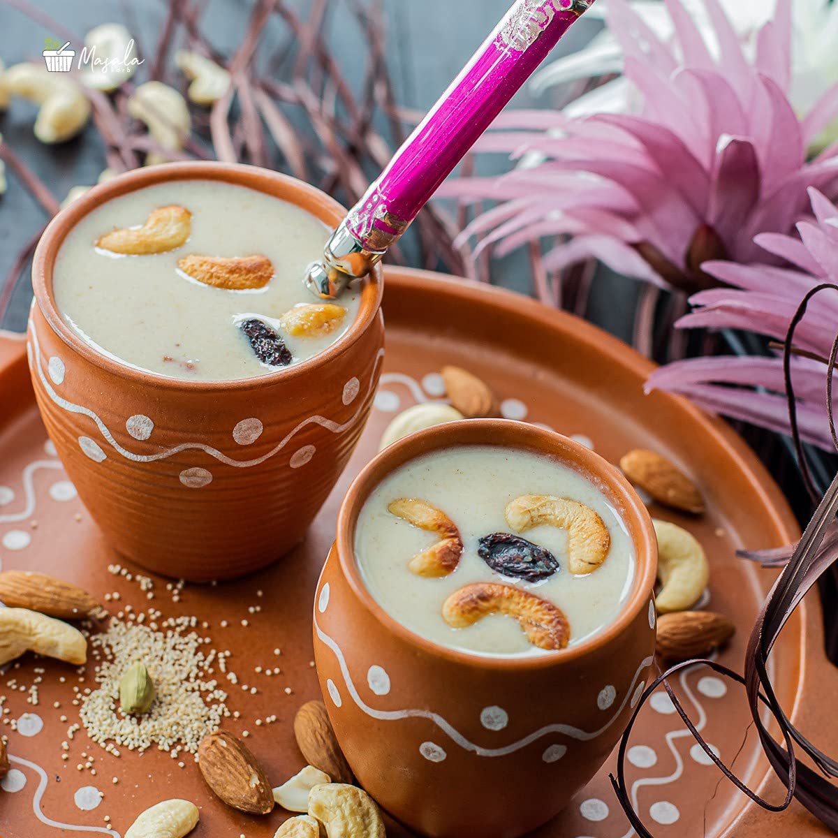 Two terracotta cups filled with a creamy beverage topped with nuts and dried fruits, on a decorative tray with pink flowers in the background.