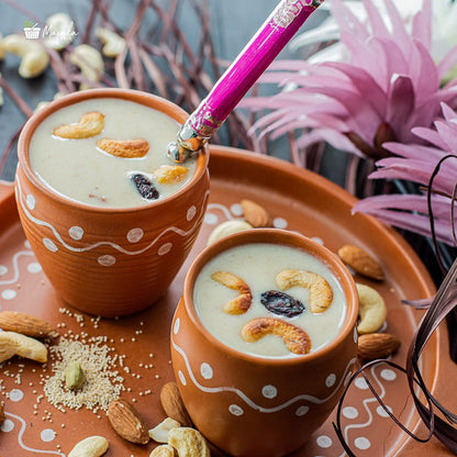 Two terracotta cups filled with a creamy beverage topped with nuts and dried fruits, on a decorative tray with pink flowers in the background.