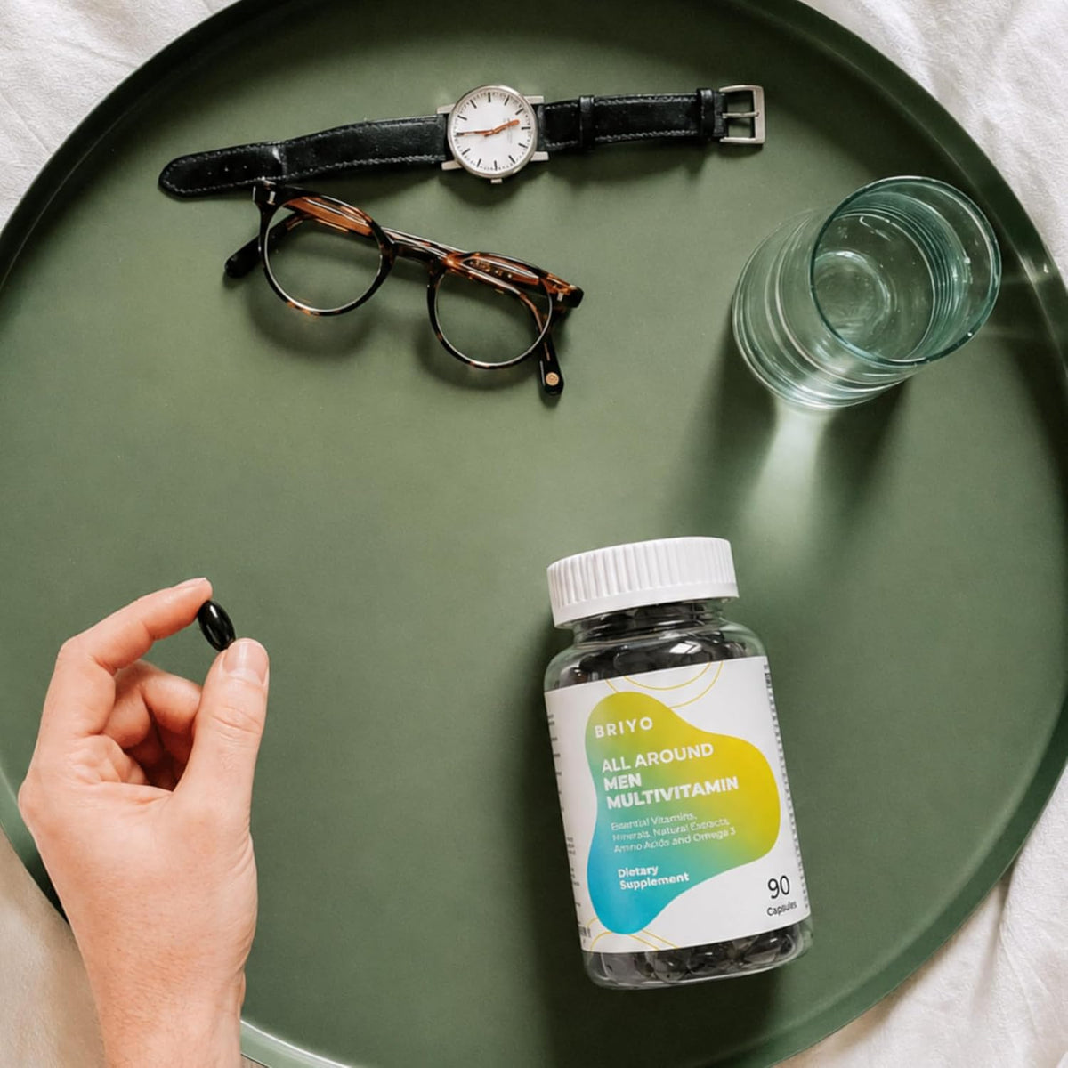 Bottle of 'All Around Men Multivitamin' on a green tray with a hand holding a supplement, glasses, watch, and glass of water.