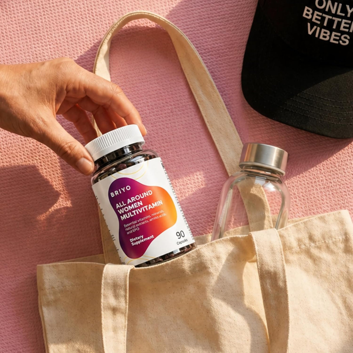 Person holding a bottle of Briyo multivitamin over a beige tote bag on a pink surface.