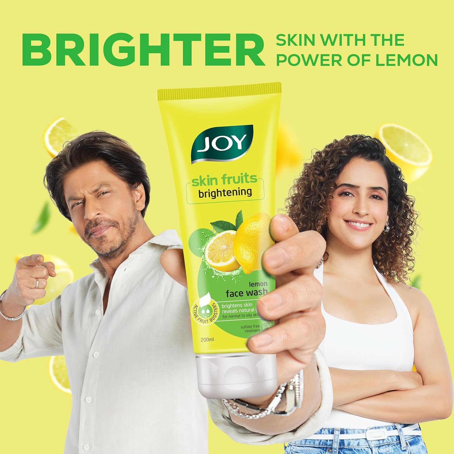 Two people holding a tube of Joy skin fruits brightening lemon face wash against a yellow background with lemons.