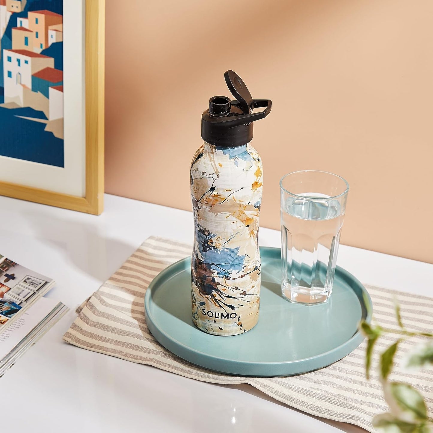 Marbled water bottle with black cap on a tray next to a glass of water on a table.