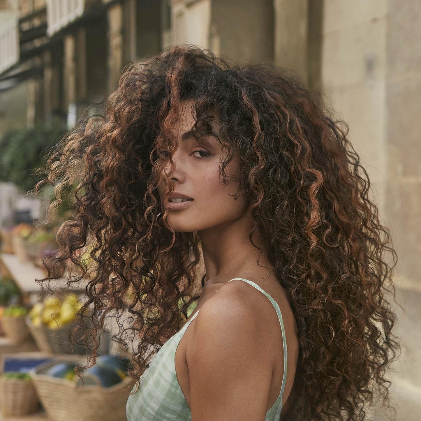 Woman with curly hair standing outdoors with a market in the background
