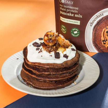 Stack of chocolate pancakes with whipped cream and banana slices on a plate, with a Cosmix pancake mix box in the background.